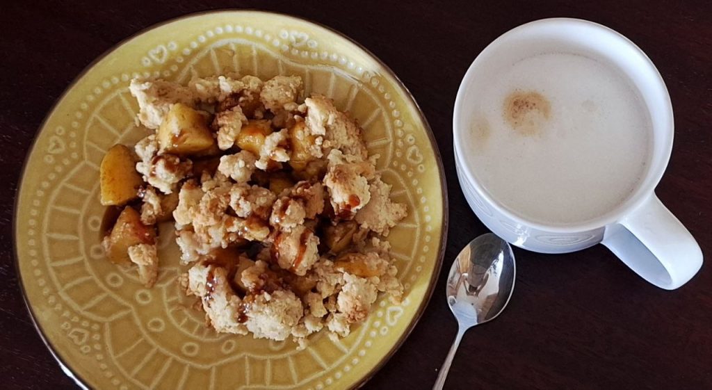 gelber Teller mit Pfirsich Crumble und daneben eine mit Milchschaum sowie ein Teelöffel auf dunkler Tischoberfläche